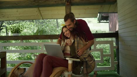 A-brunette-guy-in-a-red-T-shirt-envelops-a-girl-while-she-sits-on-an-armchair,-looks-at-a-laptop-screen-and-drinks-tea-n-the-gazebo-against-the-backdrop-of-the-forest.-The-manifestation-of-care-of-a-young-guy-is-taken-to-a-girl