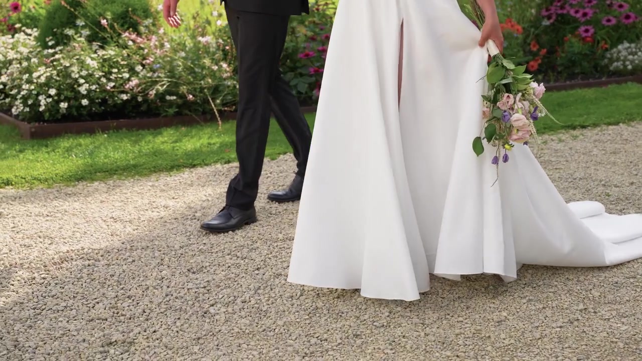 Brud i flydende hvid kjole med buket i hånden går ved siden af brudgommen på en grussti omgivet af farverige blomster i en solbeskinnet have, der fremkalder romantik og fest i filmisk stil