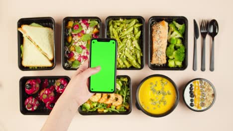 food delivery top view, take away meals in disposable containers. lunch boxes with cooked vegetarian dishes, using phone with chroma green screen. healthy diet. catering service