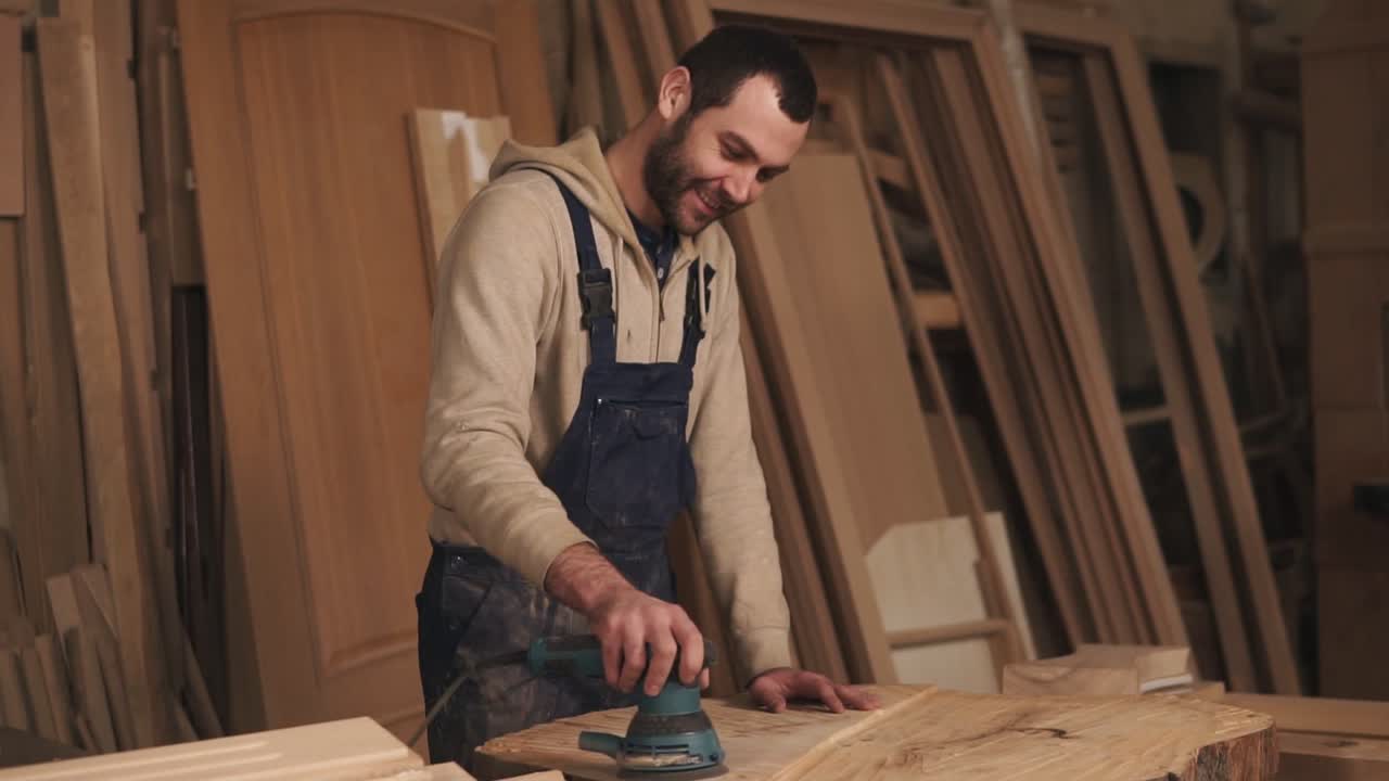 Working man is polishing a piece of wood with a machine for grinding in the hands. Wood material behind