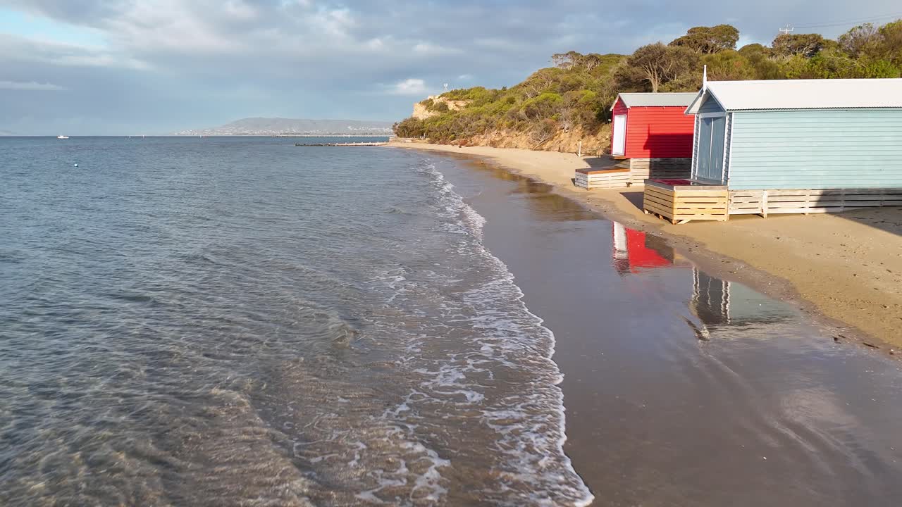 Drone glides along sandy shoreline, colorful bathing boxes, gentle waves, soft natural morning light