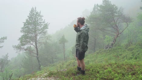 camper drinking hot coffee while standing at the campsite on a foggy morning in the forest