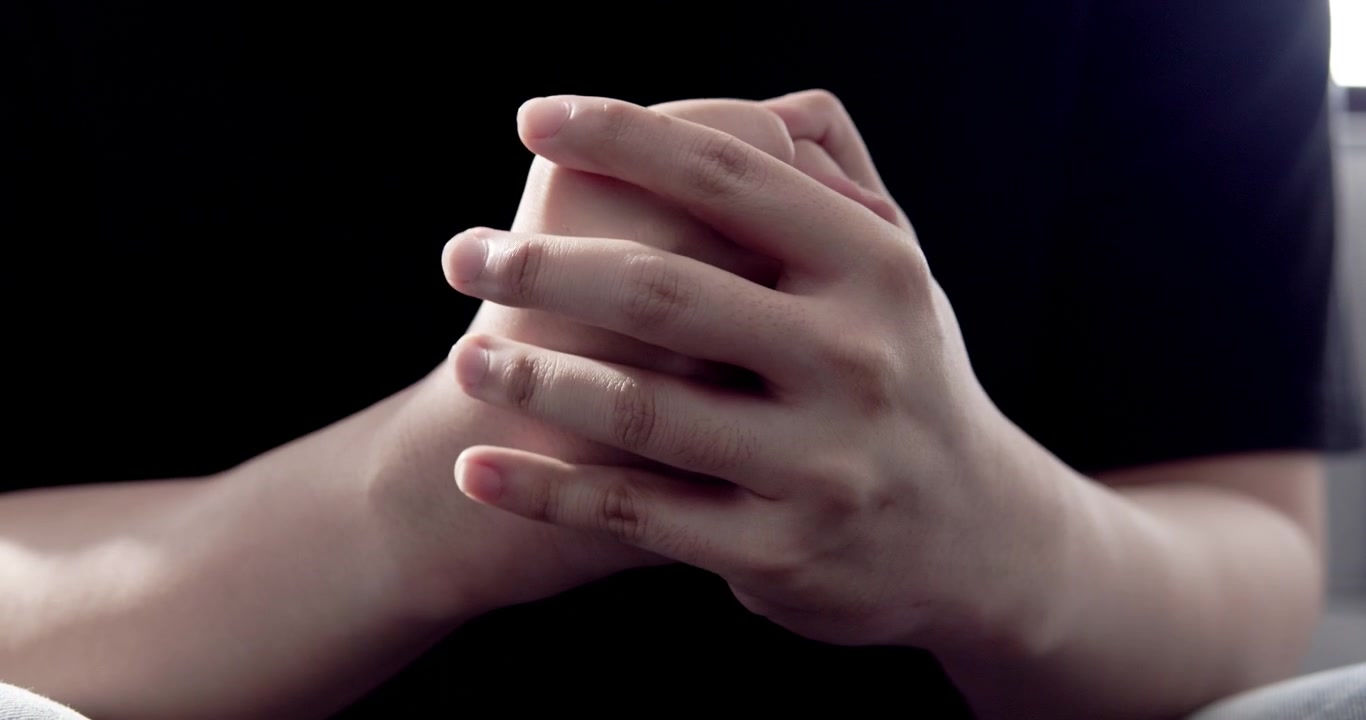 Le mani di un giovane uomo seduto su un divano nel soggiorno, strettamente congiunte per lo stress. Questa immagine trasmette profonde emozioni di tristezza e pressione.