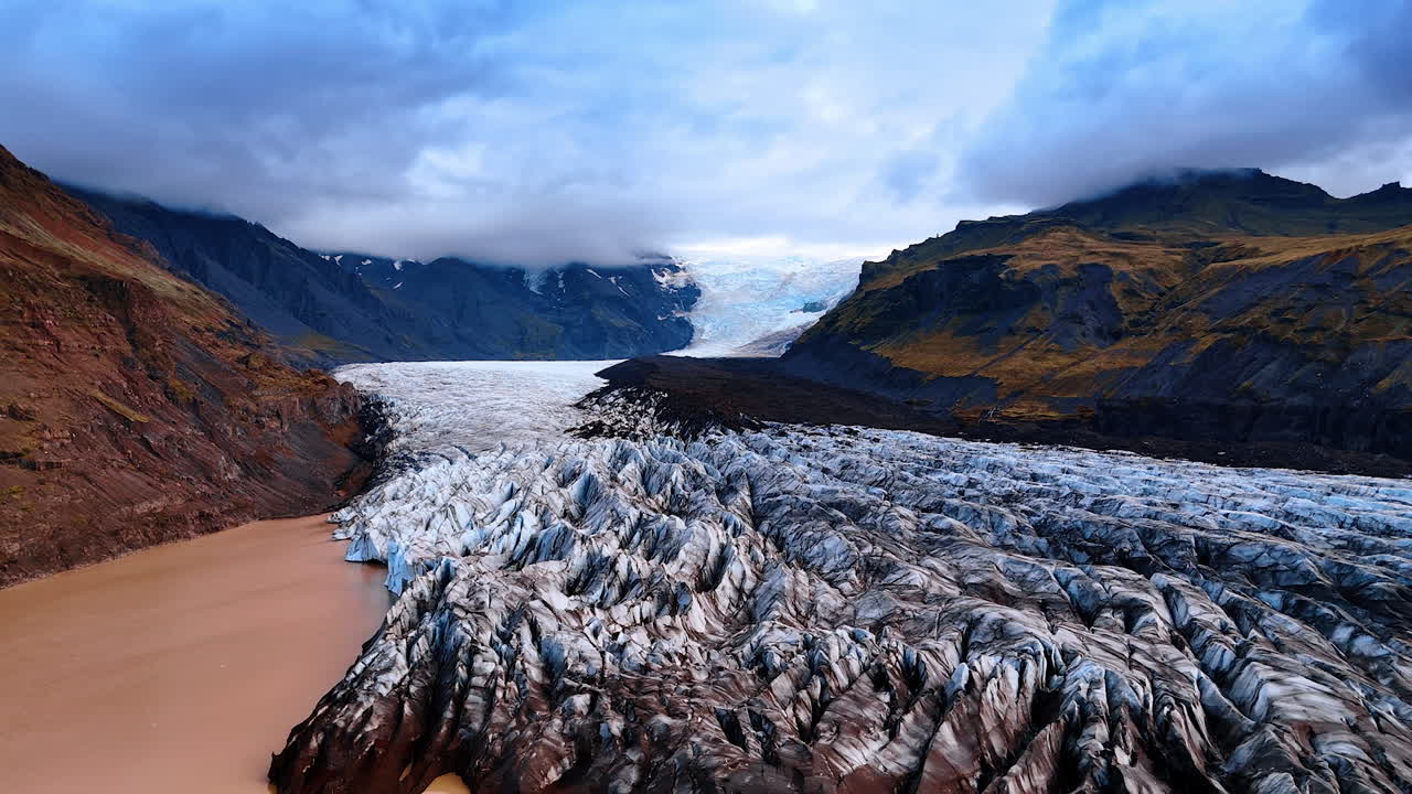 Huge glacier among the bare mountains. Heavy cloudscape hanging over the rocks hiding their peaks. Striking panorama of Iceland from top view.