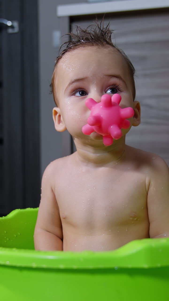 Funny boy holding a toy in his mouth. Lovely Caucasian child washing in the tub drops the toy and looks up. Vertical video