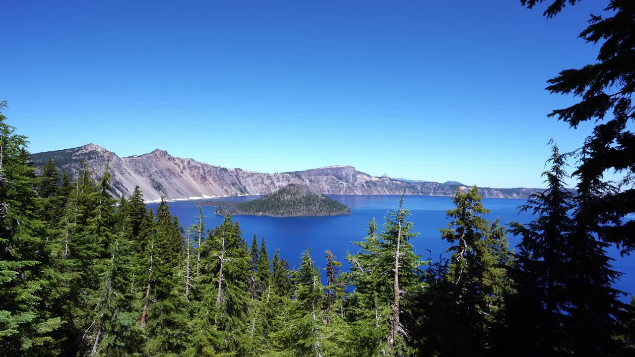 A scenic view of Crater Lake in Oregon framed by pine trees, featuring Wizard Island, blue water, and rugged mountain cliffs under a clear summer sky