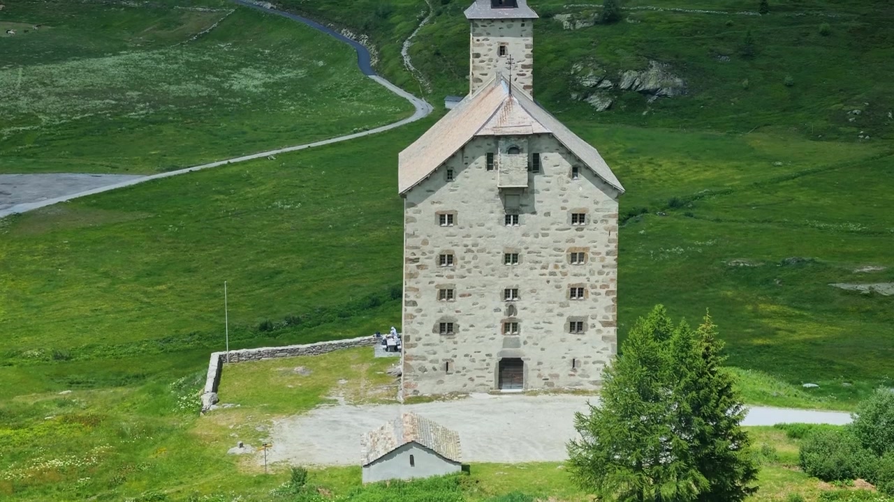 Historic Old Hospice (Alter Spittel) on Switzerland's Simplon Pass stands amid lush green meadows and stunning alpine scenery on a bright, sunny summer day