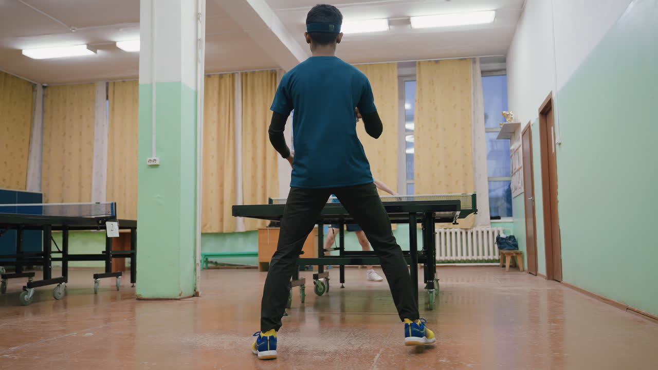 Indoor tennis tournament moment showing young player with racket ready to strike against senior opponent on polished court with yellow curtains in background