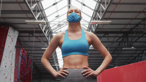 fit caucasian woman wearing face mask standing with hands on her hips in the gym