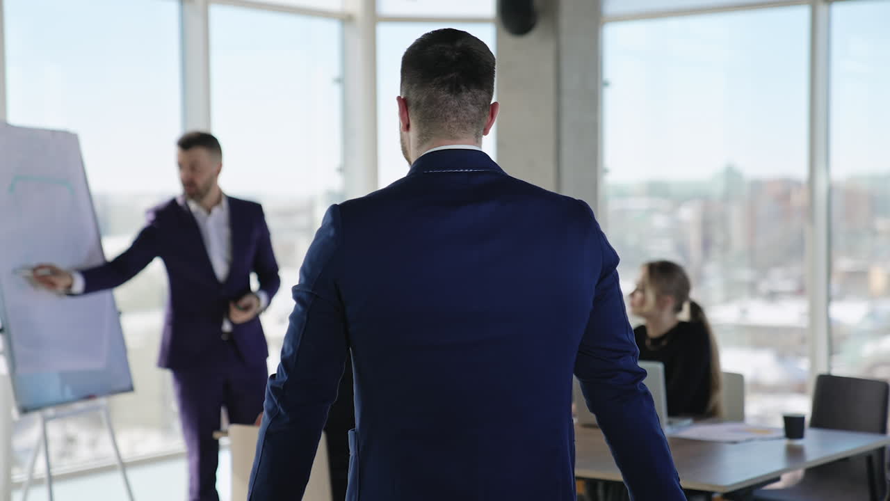 Handsome mature man in suit stands at foreground and then turns back and goes to the table. Team listening to their speaker at backdrop.