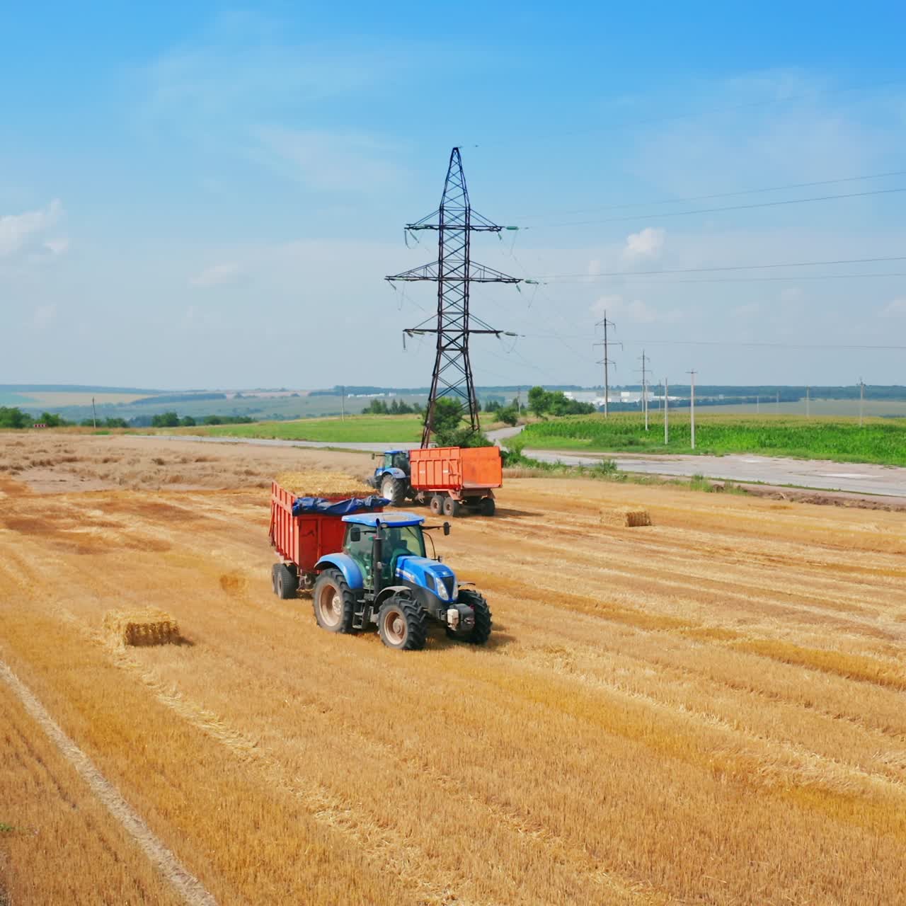 Loaded tractor full of straw leaves the field. Other agricultural machinery standing in the farmlands. Green plantations at backdrop