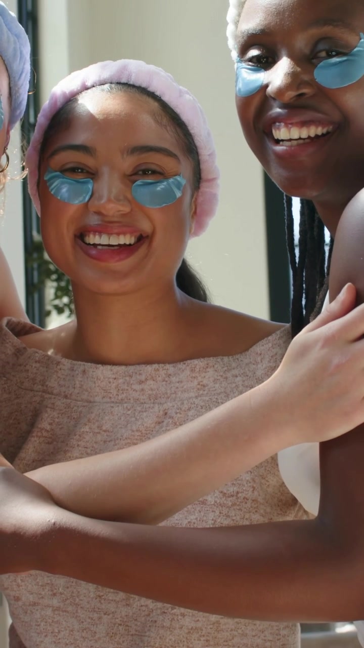 Vertical video: Laughing woman reaching and hugging other at home, wearing headbands, blue patches