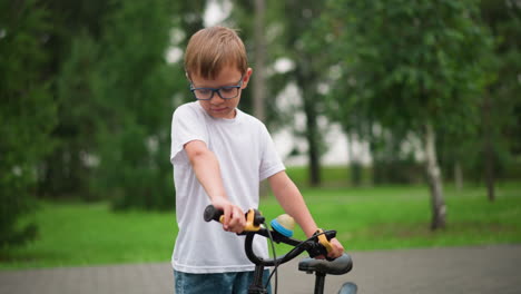 a young boy with glasses focuses on holding the brakes of his bicycle while looking down, wearing a white shirt, he is surrounded by greenery, with trees in the background