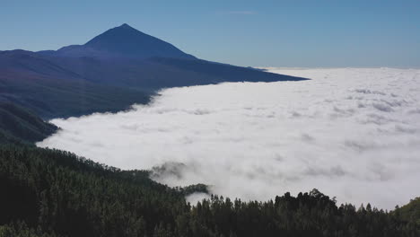 Toma-Aérea-De-Una-Vista-Desde-El-Pico-De-Teide-En-Las-Islas-Canarias-Durante-Una-Fuerte-Inversión-De-Nubes-Debajo-De-Las-Montañas-Y-Bosques-Y-Un-Cielo-Azul-Claro-Arriba