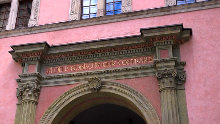Testo latino che recita SI DEUS NOBISCUM QUIS CONTRA NOS corona un arco barocco rosa con colonne in pietra a Cracovia