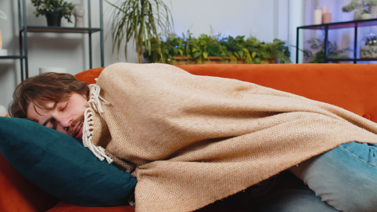 Tired young man lying down on comfortable couch taking a rest daytime nap relaxing falling asleep