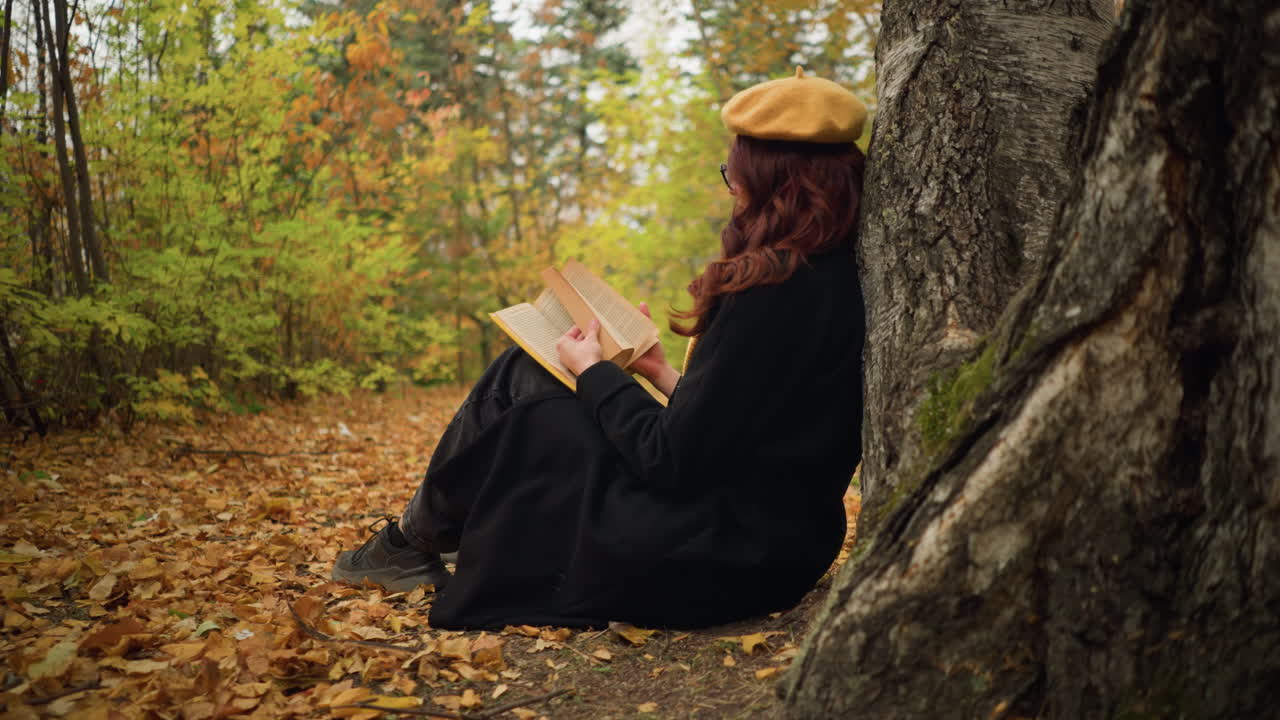 vista lateral de una mujer joven hojeando un libro en el bosque de otoño, deteniéndose con la mano en la página, sentada contra un árbol, disfruta de la lectura y la soledad mientras las hojas doradas cubren el suelo del bosque