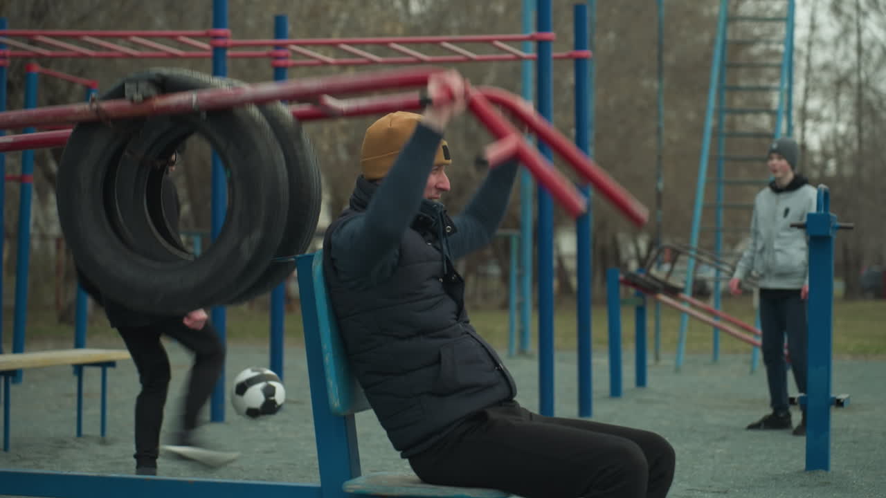 A close-up shot of a coach pausing during his workout on outdoor equipment, rubbing his hands as he rests, in the background, two boys are playing football