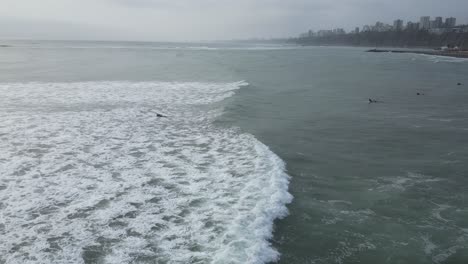 Toma-Panorámica-Aérea-Del-Mar-En-Lima-Perú-Con-Olas-Tranquilas-Y-Surfistas-Con-Tablas-De-Surf-En-El-Agua-En-Un-Día-Nublado