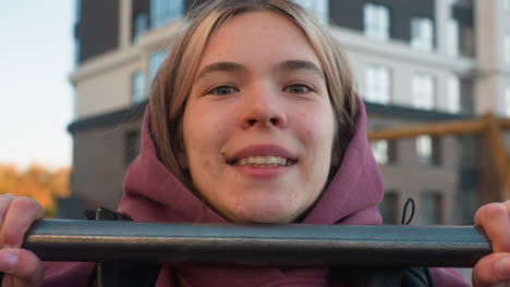 portrait of female athlete in hoodie smiling while pulling up on horizontal bar in urban outdoor gym with modern apartment backdrop, capturing exhilarating strength balance and determination