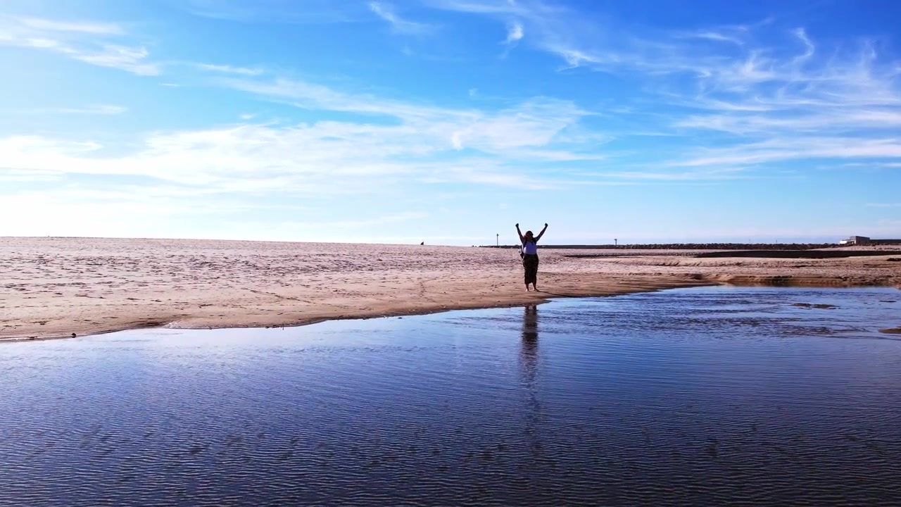 Aerial 4K shot of a person celebrating on the beach, lifting both arms towards the camera with a joyful expression, surrounded by the ocean and sandy shoreline