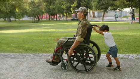 chico lindo ayudando a un padre militar discapacitado a llevar una silla de ruedas pesada