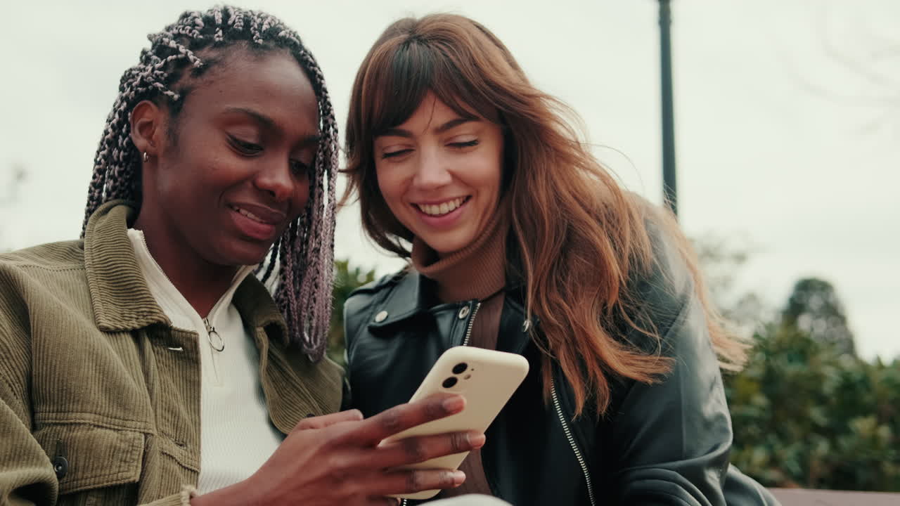 Two Young Women Friends Smiling and Talking While Looking at Smartphone