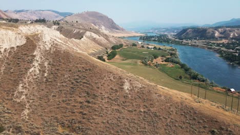 Hermosa-Vista-Aérea-Del-Campo-Verde-Y-El-Lago-Con-Montañas-Y-Cielo-Nublado-Como-Fondo