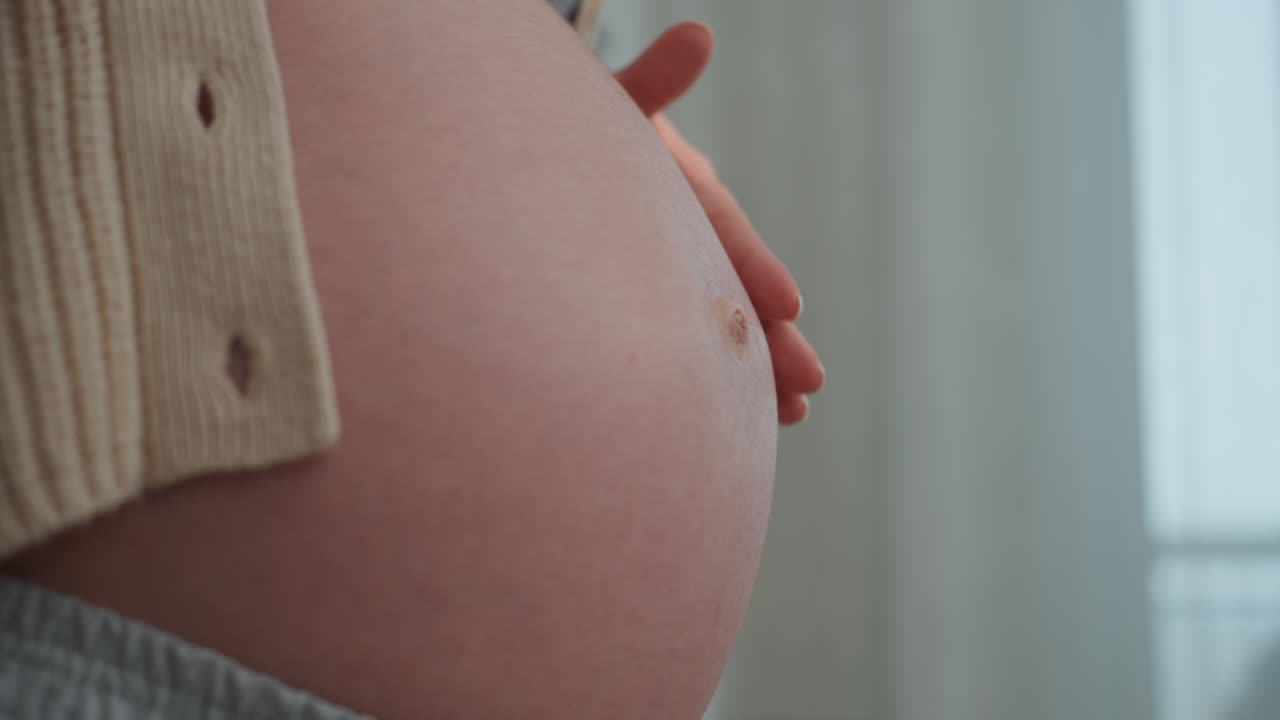 Donna caucasica incinta che accarezza la pancia vicino alla finestra, primo piano sull'ombelico e sulla pelle, movimenti delicati delle mani, cardigan tirato indietro, connessione e legame materno intimo, luce naturale soffusa, prenatale