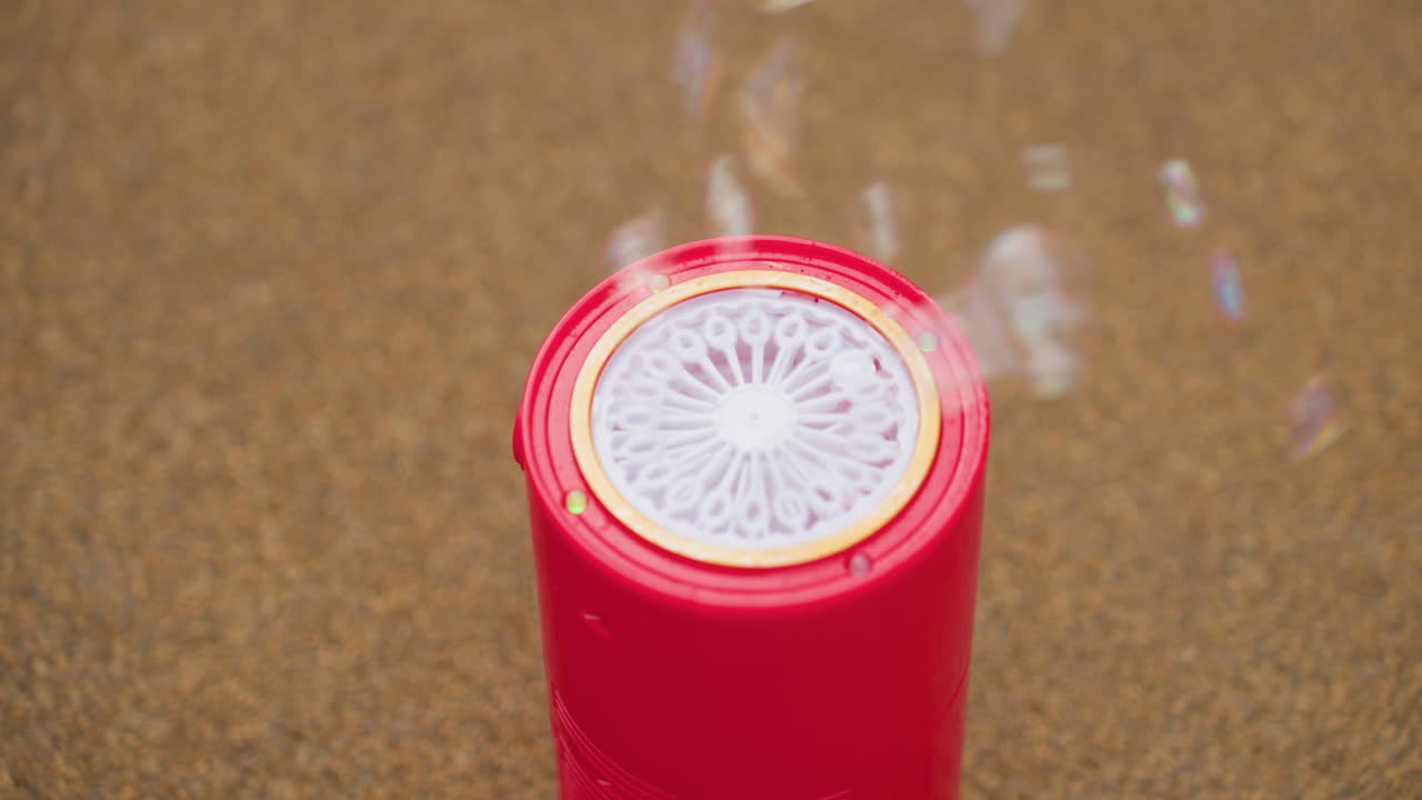 Close up red bubble machine sits on pavement releasing light soap bubbles upward, circular grille visible, soft focus ground around, playful outdoor moment capturing delicate motion and shimmer