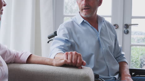 Close-Up-Of-Senior-Couple-With-Man-In-Wheelchair-Sitting-In-Lounge-At-Home-Talking-Together