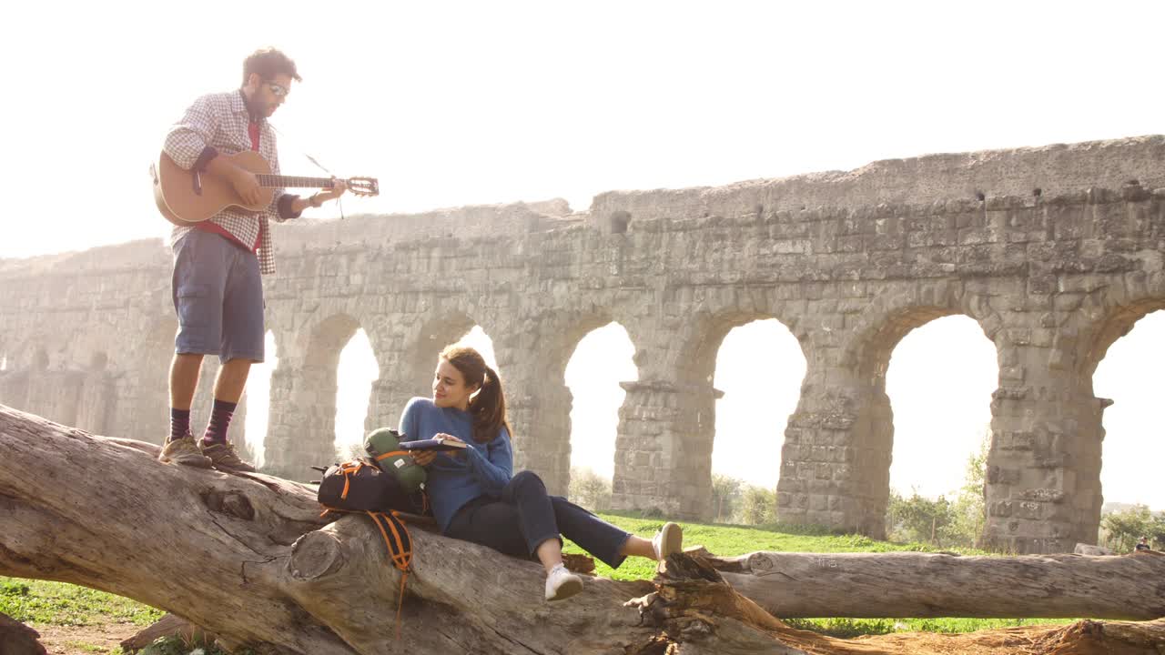 pareja feliz de mochileros turistas en un tronco de troncos tocando la guitarra cantando frente al antiguo acueducto romano ruinas en el romántico parco degli aquedotti parque en roma al amanecer trípode de cámara lenta