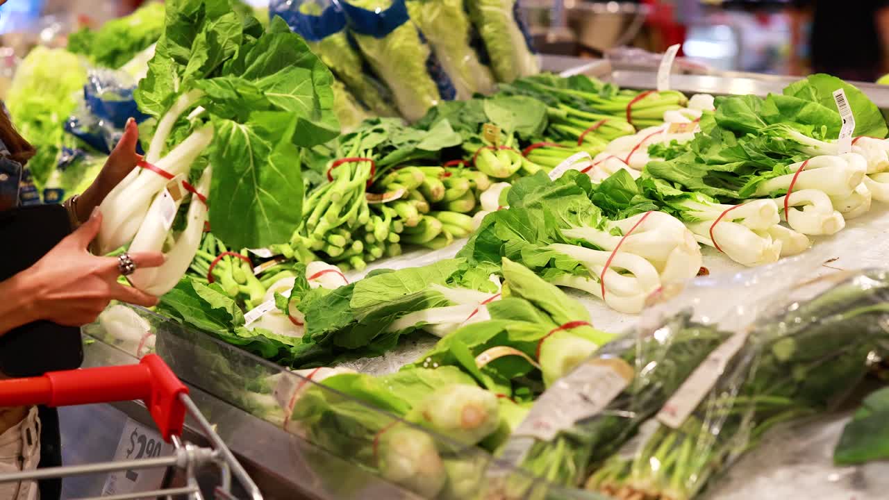 A person picks bok choy from a vibrant produce display under bright supermarket lighting