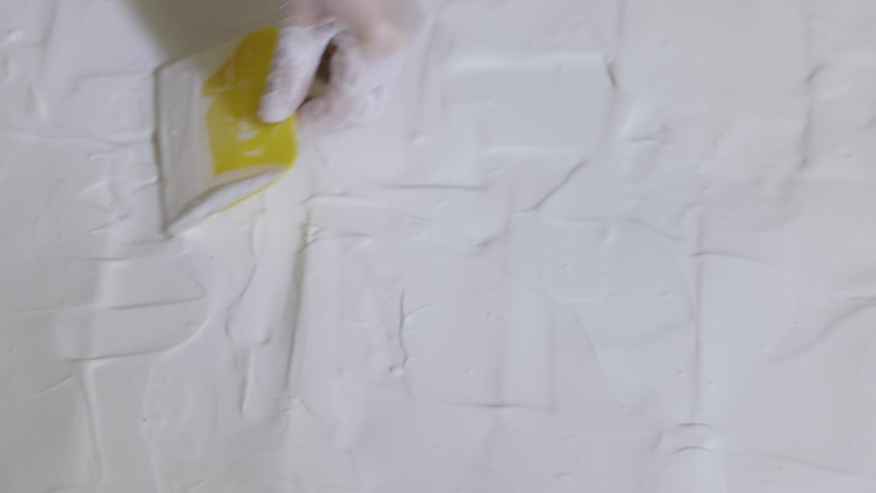 A woman's hand wearing a protective glove spreads the liquid plaster that fills the entire frame of the painting with a squeegee