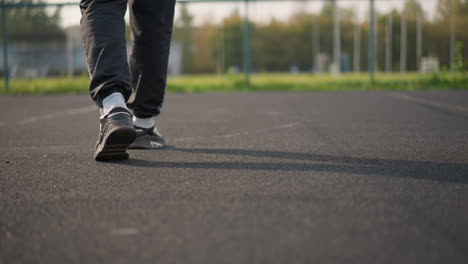 leg view of person wearing joggers and sneakers walking on court with blurred background featuring bar fence and greenery, focus on footwear and court surface in outdoor environment