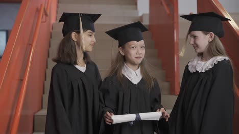 three happy preschool female students in cap and gown standing on stairs, holding diploma and talking together at the graduation ceremony