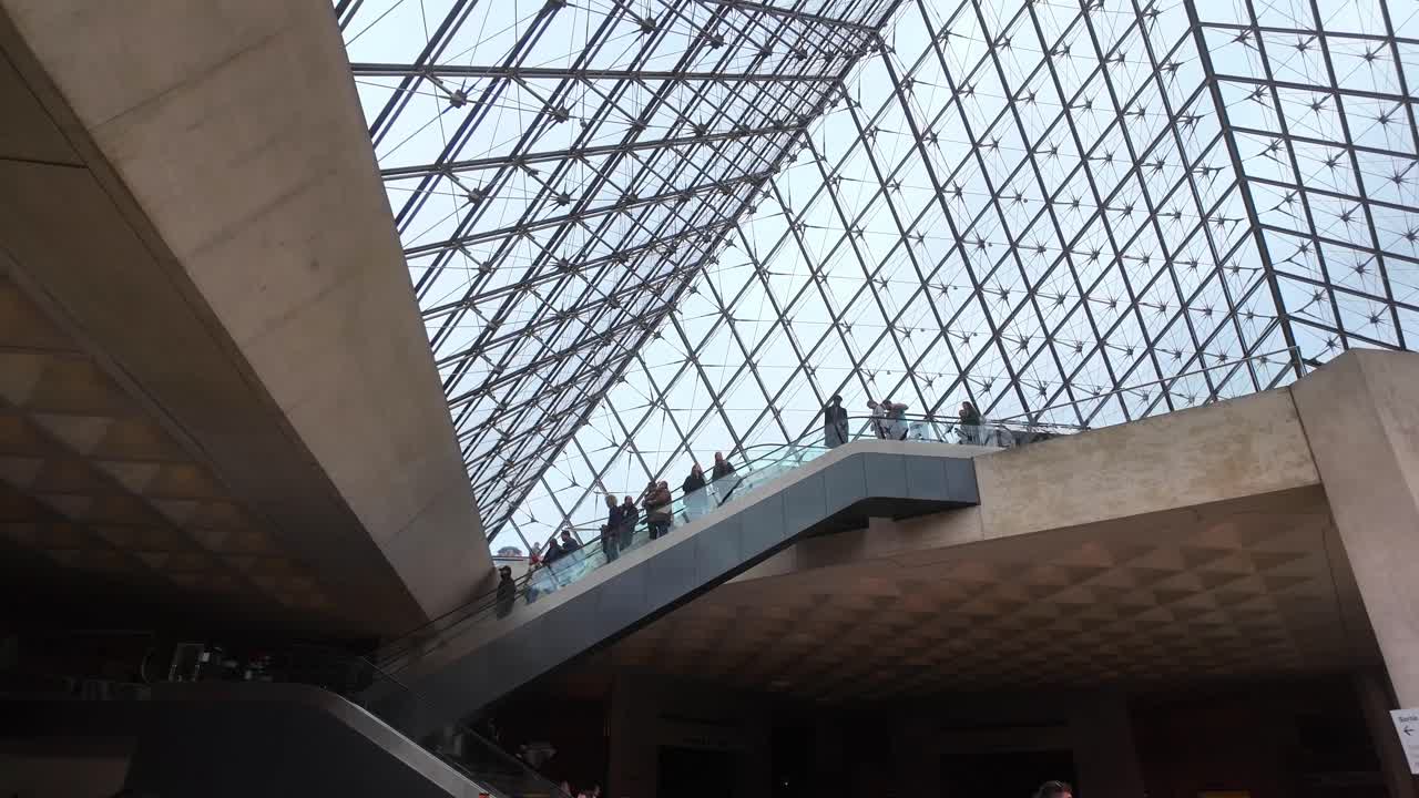 underneath view of the Louvre pyramid in daylight in Paris. People coming down the elevator beneath the entrance with interior structure visible
