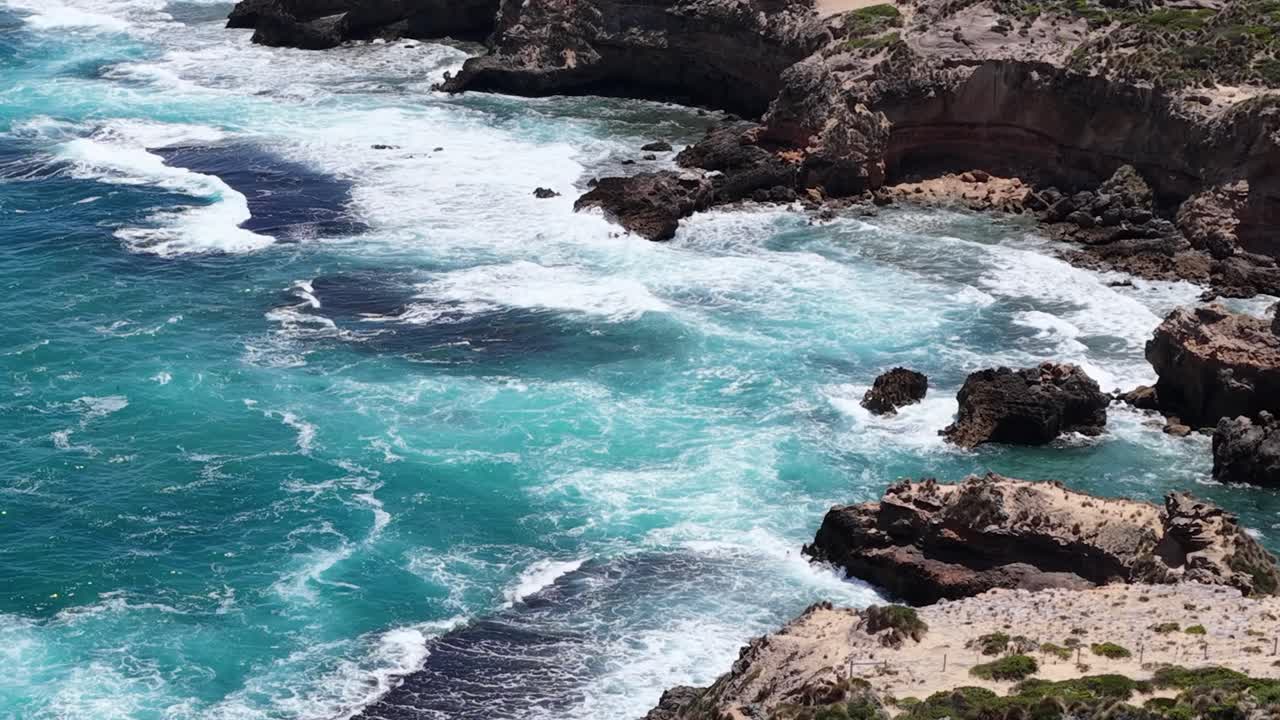 Uma foto de drone em ângulo alto captura ondas oceânicas turquesa quebrando sobre rochas costeiras acidentadas em plena luz do dia.