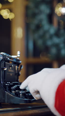 santa claus is sitting at the table and typing a letter of congratulations for the new year on the typewriter in the room on a blurred background. close-up vertical video