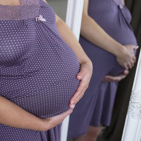 beautiful pregnant lady in a nightdress standing in front of a mirror and looking at her abdomen. future mom caresses her future child in the tummy. close up