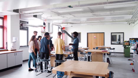 een leraar die praat met een groep middelbare schoolstudenten die rond een werkbank staan in een les over ontwerp en technologie