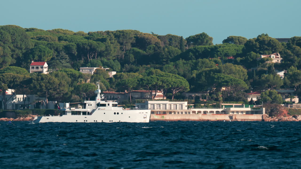 Luxury white yacht sailing near Mediterranean coastline with elegant villas surrounded by trees