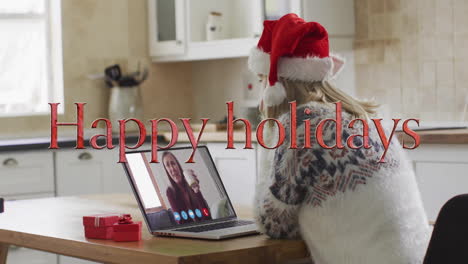 Animation-of-happy-holidays-text-over-caucasian-woman-on-laptop-screen-and-christmas-decorations