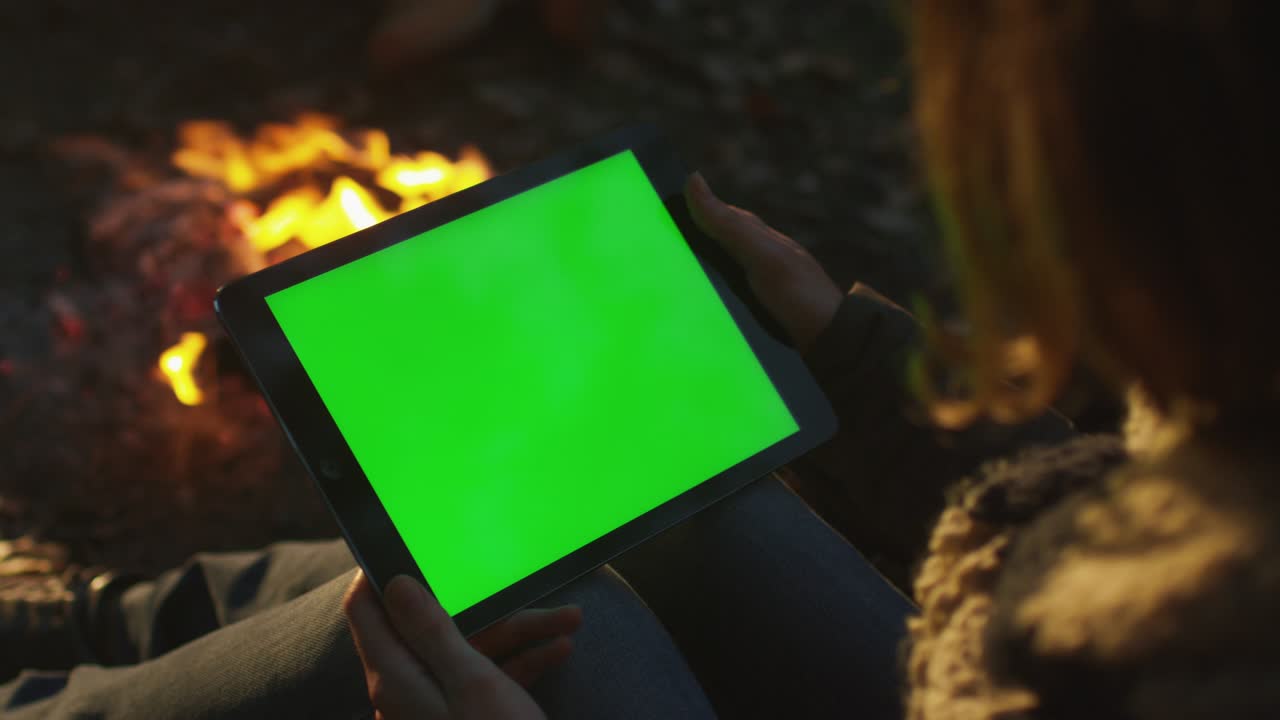 Girl is holding a tablet in landscape mode with green screen mock-up next to a campfire in the evening.