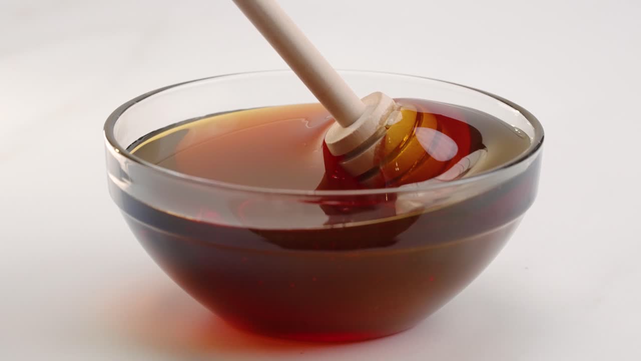 Miele in ciotola mescolato con un bastoncino a nido d'ape al rallentatore su sfondo bianco da studio in 4k.