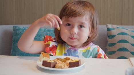 little child girl eating raisins, sitting on couch and pick up dried grapes from plate with snackes and cakes