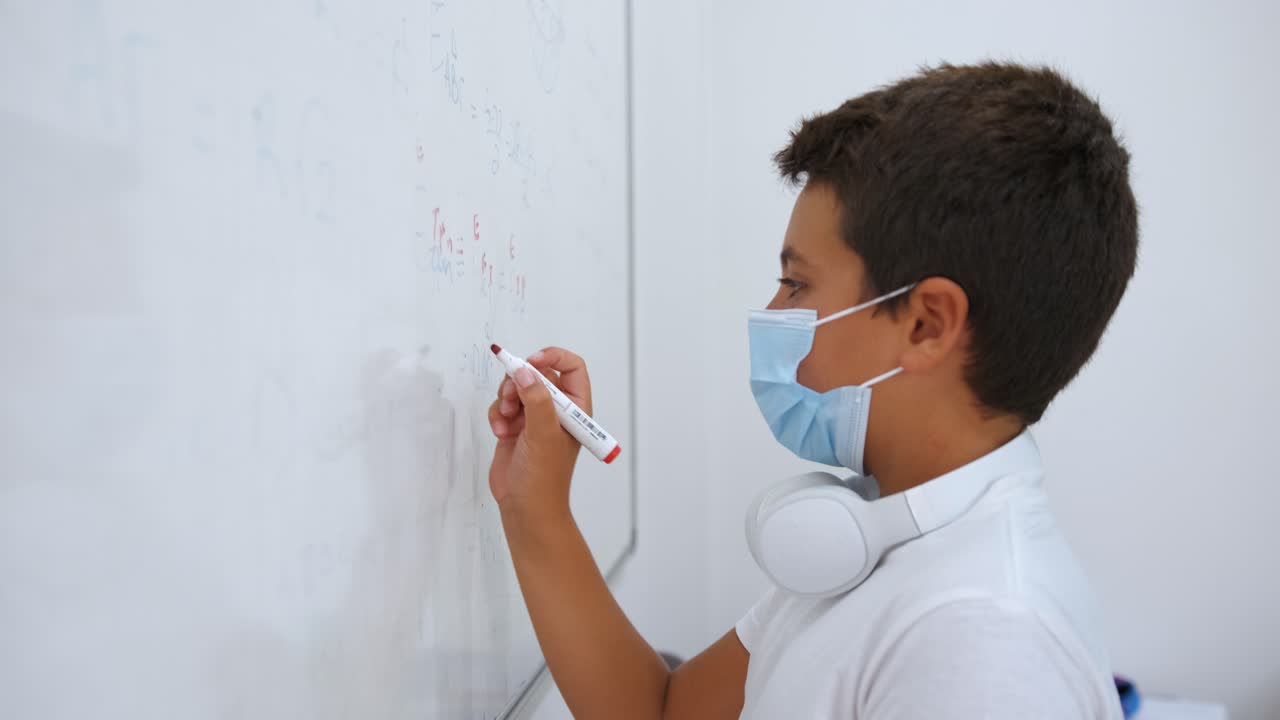niño de escuela con mascarilla respondiendo preguntas de matemáticas en una pizarra blanca con marcador rojo