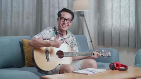 asian man composer with headphones smiling and showing thumbs up gesture to camera while playing guitar at home