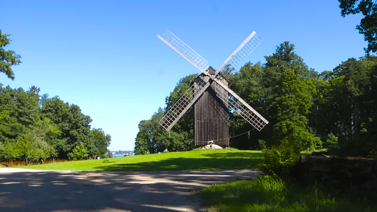 Traditional old and large wooden windmill or milling grain structure with large paddles in a sunny summer countryside seaside garden with gravel roads and green mowed grass around it. Blue sky visible