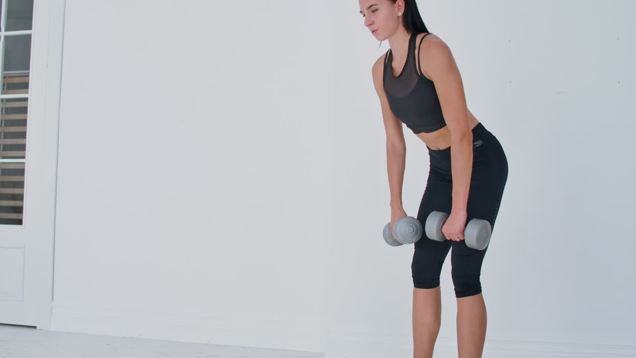 uma jovem atleta em um apartamento brilhante realiza um levantamento de peso com halteres em casa fazendo inclinações para a frente para fortalecer os músculos das coxas